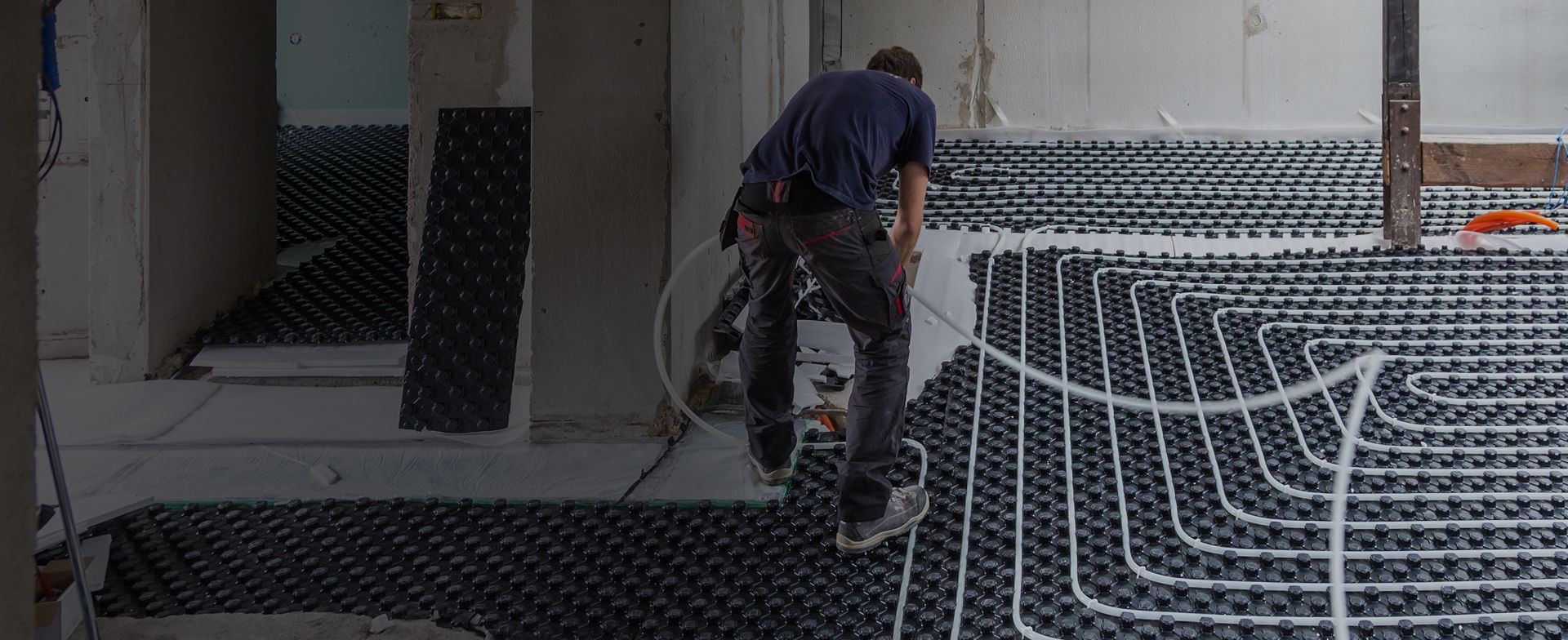 Ein Arbeiter verlegt Fußbodenheizungsrohre auf einem schwarzen Kunststoffgitter in einem renovierten Raum und bereitet den Bodenbelag vor.