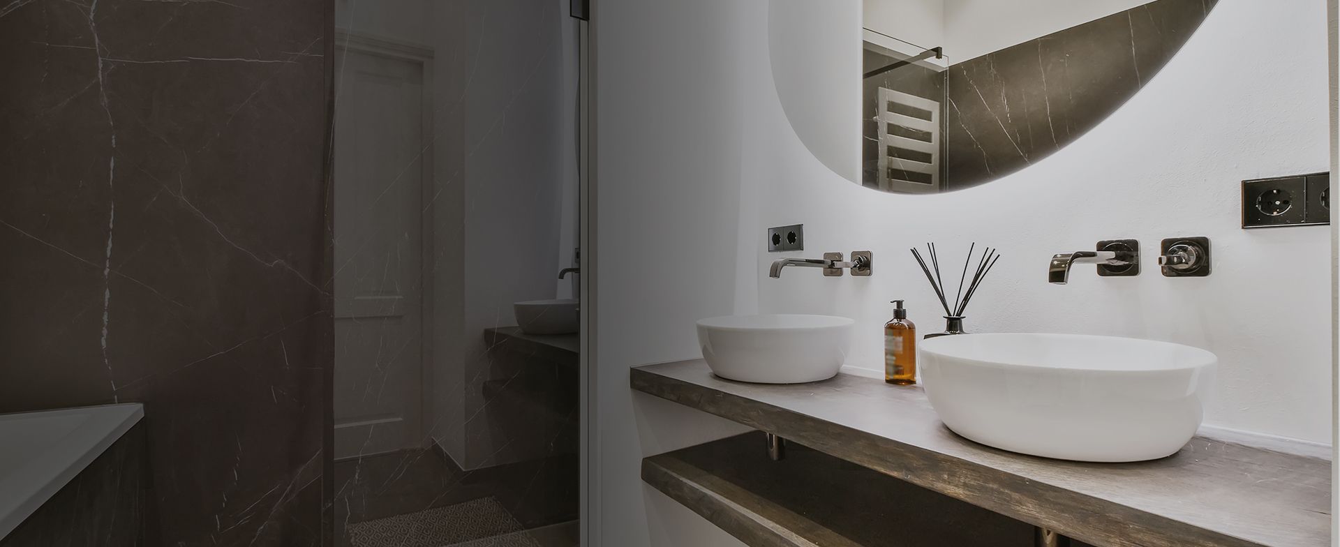 Modernes Badezimmer mit zwei weißen ovalen Waschbecken, eleganten Armaturen, einer bernsteinfarbenen Flasche und dekorativen Rohrstäben vor dunklen Marmorakzenten.